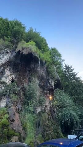 A video shows a cliff with a small waterfall and lush green vegetation. The cliff face is rocky and moss-covered, with water trickling down it in several places. Trees and other plants grow on top of and around the cliff. A single streetlight is visible on the cliff face. In the foreground, the hoods of two cars are visible, one dark-colored and one blue. The sky is clear and blue, suggesting it is daytime. The video is brief and appears to be taken from a stationary viewpoint.
