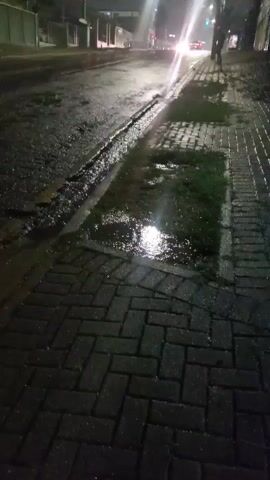 The video presents a nighttime street scene in Curitiba, Brazil, characterized by wet surfaces and artificial illumination. The camera is positioned low on a brick-paved sidewalk, looking down a street. The street, made of asphalt, is thoroughly wet, suggesting recent or ongoing rain, with prominent puddles reflecting the bright light sources. A distinct gutter or channel filled with water runs along the curb.

A powerful streetlight above casts a brilliant beam downwards, causing a significant lens flare across the frame and brightly illuminating a large puddle on the sidewalk. In the distance, a vehicle with intense headlights approaches, quickly drives past the camera's view, and then recedes, its lights creating dynamic reflections on the wet asphalt. A single person is visible walking on the far side of the wet sidewalk in the distance, their silhouette partially defined against the distant lights. Buildings and fences line the street in the background, mostly obscured by the darkness. The scene conveys a quiet, damp urban atmosphere at night. No visible text is present in the video.