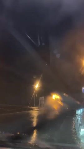 This is a night time video of a flooded road. It is raining heavily. A white truck is driving on the road and splashing water. The road is illuminated by street lights and the headlights of the truck. The water on the road reflects the lights.