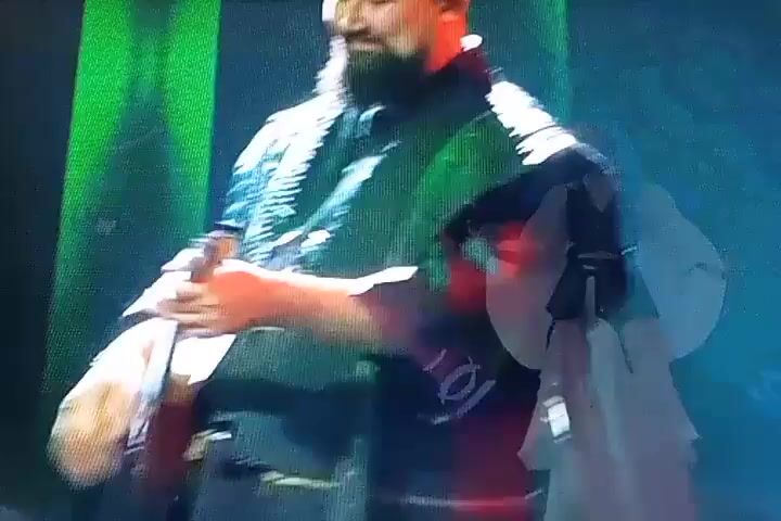 The video shows a performance on a stage. In the first shot, a man with a beard is adjusting something on his chest, possibly a microphone or an instrument. Green lights illuminate the scene. The next shot shows the same man, now singing into a microphone with blue lights affecting the scene. The third and fourth shots show a woman in traditional costume, possibly performing a dance or playing an instrument. She is holding a microphone and gestures with her hands. The background features abstract blue and purple lights. The final shot returns to the man, who is now more clearly singing into a microphone and appears to be addressing the audience. The lighting shifts between green and blue.