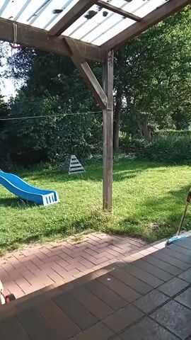 The video shows a backyard scene. In the foreground, there is a patio with brick pavers. A wooden pergola with a translucent roof covers part of the patio. Beyond the patio is a grassy lawn. On the left side of the lawn, there is a blue plastic slide for children. In the background, there are trees and a small white pyramid-shaped structure with black stripes. The sun appears to be shining, casting shadows. The overall impression is of a bright, outdoor setting, likely a residential backyard.