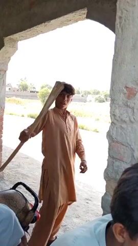 A group of young boys are playing marbles outdoors. In the first few seconds, a boy in a tan shalwar kameez stands under an overhang and gestures towards the camera. He then jumps forward, making a face and holding a stick. The scene shifts to show two boys in blue and white shalwar kameez. The boy in blue is standing on a small object with his hands covering his ears, appearing to be playing a game. A few other boys are visible in the background. The video continues to show the boys playing marbles with several marbles visible on the ground.