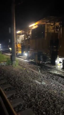 At night, a large yellow train-like machine with the text UNIMAT 08 visible on it is operating on railroad tracks. The machine has numerous mechanical components and is illuminated by bright lights. It appears to be actively working on the tracks, with tines or probes extending downwards into the ballast. The scene is dark, with only the machine and its lights providing illumination.