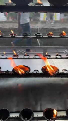 The video shows a rack of oil lamps with flames burning. In the background, there are people walking. The lamps are arranged in multiple rows, and some of the flames are quite large and flickering. The environment appears to be outdoors, with some greenery visible in the background. The lighting is dim, with the flames of the lamps being the primary light source. The camera is positioned close to the lamps, focusing on the flames and the metal rack.