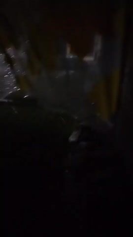 The video shows a dark, outdoor scene, possibly at night, with rain falling. The camera is held close to the ground, looking upwards. In the foreground, there is a dark, blurry surface with some reflective elements, suggesting it could be a wet ground or possibly the inside of a transparent object like an umbrella. As the camera moves, it captures glimpses of a yard with grass and a tree. The background is very dark and indistinct, with some blurry lights visible in the distance, possibly from buildings. The overall impression is of a brief, handheld recording during a rainy night.