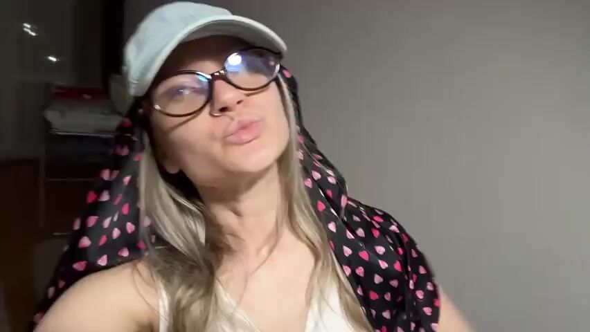 A fair-skinned woman with blonde hair, wearing a light-blue cap, round glasses, a nose ring, a white tank top, and a black scarf with pink hearts, poses for a selfie. She purses her lips and holds up her right hand, showing off red nail polish. The background includes a couch, pillows, and a wooden floor. The lighting is somewhat dim.