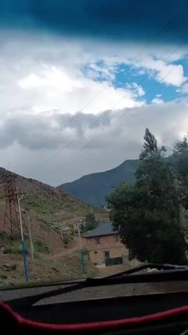 The video is filmed from inside a vehicle looking out the windshield. The camera is tilted upwards, showing a cloudy sky with patches of blue. In the middle ground, there are rolling hills covered in green vegetation and scattered houses. A road curves through the landscape. In the foreground, a dashboard with red trim is visible. A decorative object, possibly an air freshener or charm, hangs from the rearview mirror. The overall impression is a scenic drive through a rural, mountainous area.