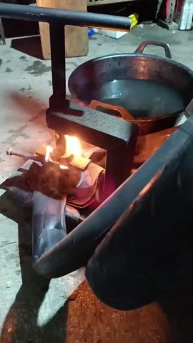 The video shows a close-up view of a tire repair process. A bicycle inner tube is being heated by an open flame from a makeshift stove. A metal clamp is pressing down on the inner tube, which is placed on a metal stand. A large black wok filled with liquid is visible in the background. The scene appears to be taking place outdoors or in a workshop, with a concrete floor and some shelving units in the background. The lighting is dim, with the flame and the hot metal object being the primary light sources. The overall impression is one of manual labor and resourcefulness.