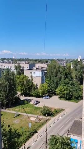The video shows an aerial view of a residential area on a sunny day. The camera pans over apartment buildings surrounded by trees. There are cars parked on the street and in a courtyard. The sky is clear blue with a few white clouds. A thin wire stretches vertically from the bottom of the frame towards the top.