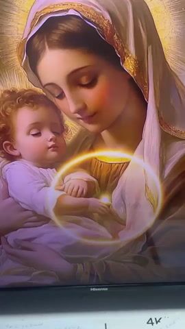 The video displays a religious artwork depicting the Virgin Mary holding the infant Jesus. The image is illuminated by a radiant golden halo surrounding Mary's head and a glowing orb encircling her and Jesus. The scene appears to be displayed on a screen, with a "Hisense" logo visible at the bottom, suggesting it's a digital display. The lighting is soft and ethereal, with a focus on the gentle expressions of Mary and Jesus.