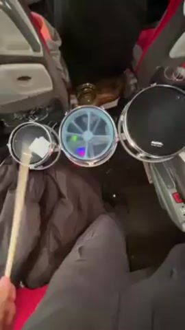 The video shows a person seated on an airplane, playing a miniature drum set. The drum set consists of three small drums placed side by side. The drum on the left is covered with a white drumhead, the middle drum has a clear head with visible blue and green lights inside, and the drum on the right has a black drumhead. The drums are arranged on a surface covered with a black padded jacket. The person is holding a drumstick and striking the drums. The airplane seats, covered in a red and gray fabric, are visible on either side, and passengers can be seen moving in the aisle. The video is taken from a first-person perspective.