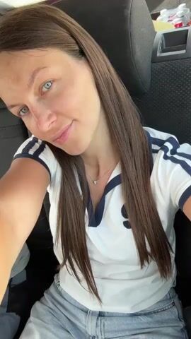 A woman with long brown hair and blue eyes sits in the backseat of a car. She is wearing a white shirt with blue stripes on the sleeves and a small silver necklace. She is smiling and looking at the camera. The car is moving and the background is blurred.