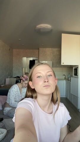 A young woman with light skin and long blonde hair stands in front of the camera, applying makeup to her face with a large, black, dome-shaped brush. She is wearing a light pink t-shirt. In her other hand, she holds a small makeup sponge applicator. The room appears to be a small studio apartment with a kitchen in the background, complete with white cabinets, a small refrigerator, and a microwave. To the left, a bed is partially visible, and another person is lying in it, covered by blankets. The overall lighting is soft, and the focus is on the woman and her makeup application.