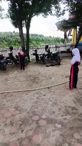 The video shows three young girls dressed in black tracksuits with red stripes and white headscarves playing a game. The girls are barefoot. They are standing in a cleared area with a patterned brick surface. A white rope lies on the ground between two of the girls. There are several parked motorbikes behind the girls and a white fence beyond the motorbikes. Behind the fence is a river or body of water with trees and foliage on the opposite bank. The sky is overcast. There are trees on the left and right sides of the frame. The girl on the left is holding a small, dark object, possibly a ball. The girls are taking turns kicking the object over the rope.