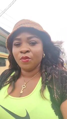 A woman is the main subject of this selfie-style video. She has brown skin, dark hair, and is wearing a brown patterned bucket hat, a neon green Nike tank top, a black crocheted shrug, and a gold key necklace. She also has gold hoop earrings and dark red lipstick. The camera is held at arm’s length, capturing her face and upper body. The background shows a busy street in what appears to be a residential or commercial area. There are buildings, cars, and other people visible. She is looking directly at the camera with a serious expression, and her hair is being blown by the wind.