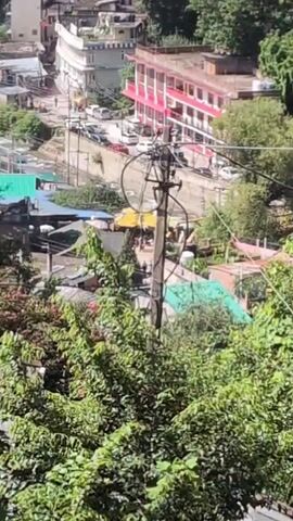 The video opens with a high-angle, slightly shaky view of a hillside town. Lush green trees in the foreground partially obscure the view. The town is built on a slope, with buildings clustered together. In the middle ground, a street is visible with parked cars and a few people walking. A utility pole with wires stands prominently. The buildings are varied in color and style, with some appearing to be residences and others possibly commercial establishments or hotels. Many buildings have red-tiled roofs. Further in the background, more buildings extend up the hillside, interspersed with trees. The sky is blue with some scattered white clouds. The camera slowly pans left and slightly upwards, revealing more of the town and the surrounding mountainous landscape. In the distance, a prominent mountain peak is visible. The video also briefly shows a window frame on the left side. The overall impression is of a bustling town nestled within a scenic mountainous region.