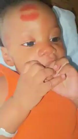 Here's a description of the video:

The video shows a baby lying on a blue and white patterned pillow. The baby has dark skin, dark hair, and is wearing an orange sleeveless shirt. There's a red lipstick kiss mark on the baby's forehead. The baby opens their eyes, looks at the camera, and puts their fingers in their mouth. The baby is also wearing a silver bracelet. The background is blurry and shows what appears to be a wooden surface.