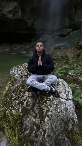 The video opens with a low-angle shot of a person sitting cross-legged on a mossy rock in front of a waterfall. The person is wearing a dark hoodie, light gray pants, and dark shoes with white accents. Their hands are pressed together in a prayer-like position. The waterfall is tall and narrow, cascading down a rocky cliff face into a pool of water. The surrounding environment consists of lush greenery and moss-covered rocks. As the video progresses, the camera angle tilts upwards, focusing on the waterfall and the cliff face. The sky is visible at the top of the frame, and there are trees lining the edges of the cliff.