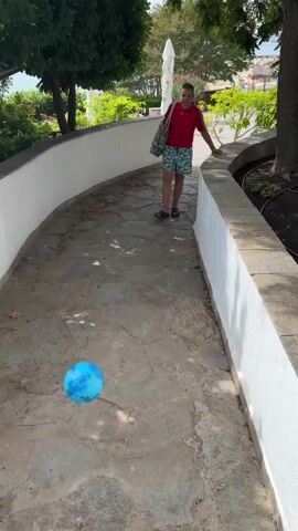 A boy in a red shirt and patterned shorts stands on a stone pathway with a blue ball at his feet. He kicks the ball, which rolls away. The pathway is lined with a white wall and trees. The boy walks after the ball. The camera pans down to show some stairs.