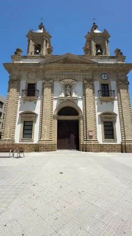 This is a vertical, eye-level shot of a large church facade on a bright, sunny day. The building is made of stone with white accents, and features two bell towers with domes at the top, each topped with a cross. In the center of the facade, above the large, dark wooden double doors, is a niche with a statue of a saint. Above the statue is an arched window, and a clock face is visible on the right side of the facade. There are balconies on either side of the central section. The ground in front of the church is paved with square tiles, and a couple of benches are visible to the left. A modern building can be seen to the far left of the frame. The sky is a clear, vibrant blue, indicating fair weather. The sun casts strong shadows, suggesting it is mid-day. The scene appears to be a public square in front of a place of worship in Cádiz, Spain. There are no people visible in the shot. There is a small pink sign on the right side of the building, but no text is legible.