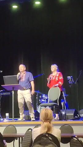 Two people are on a stage with a black curtain backdrop. A man in a blue shirt with red and blue embroidery and khaki pants stands to the left, holding a microphone and looking at a laptop on a podium. A woman in a red shirt with red and yellow embroidery stands to the right, holding a microphone and looking forward. A chair is behind her. A drum set is visible behind them. In the foreground, the back of a woman with blonde hair tied in a ponytail is visible, seated at a table facing the stage.