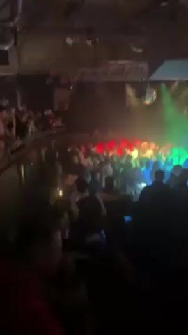 The video shows a crowded indoor venue, likely a nightclub, with many people on a dance floor. The lighting is dynamic and colorful, featuring green, blue, and red lights, along with disco balls that reflect light around the space. Above the crowd, there is a metal truss system holding lighting and sound equipment. The video is taken from a slightly elevated position, giving a view over a portion of the crowd. People are moving and interacting with each other. The overall atmosphere suggests a lively, energetic environment.