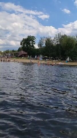 The video shows a body of water, likely a lake or sea, with a beach in the background. The sky is blue with scattered white clouds. The beach area is populated with many people sunbathing, swimming, and relaxing under umbrellas. There are some buildings, trees, and beach chairs visible on the shore. The water is rippling gently. The scene appears to be a warm, sunny day at a popular recreational area.