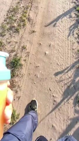 The video shows a person's hand holding a GymBeam shaker bottle filled with a light-colored liquid and ice. The person is wearing dark blue pants and black shoes and is walking on a dirt path next to a cornfield and a wheat field. The sky is clear and blue. The camera is positioned at a downward angle, looking from the person's perspective.