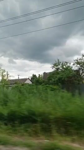 The video begins with a view of a rural area under a cloudy sky. Tall green grass and trees line the side of a dirt road. In the distance, several houses with green roofs can be seen. Overhead, power lines crisscross the sky. The camera then pans to the right, showing more houses and trees. A blue car is seen driving on the road, followed by a black SUV. The video continues to pan right, revealing more of the rural landscape. The scene is one of a quiet, possibly underdeveloped, residential area with a focus on natural elements and basic infrastructure.