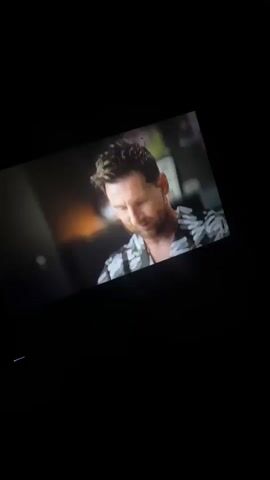 The video shows a shot on a television screen of Lionel Messi. Messi is wearing a black and white patterned shirt. A crew member stands in the background, out of focus. A clapperboard is held in front of Messi, the words "MESSI" "1/1" are visible.  The scene is dark, the video is shot inside a home theater or entertainment room.
