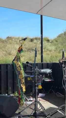 The video shows a black stage set up outdoors against a backdrop of grassy dunes under a blue sky. A white canopy covers part of the stage. A black speaker sits on the left side of the stage. A microphone on a stand is draped with a brightly colored scarf with yellow flower patterns. Other microphone stands and cables clutter the space. Musical instruments are visible, including a small drum kit and a xylophone. The stage appears to be prepped for a performance, but no one is present in the shot.