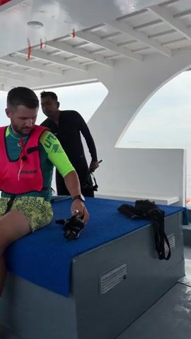 The video shows a group of people on a boat, preparing for a snorkeling or diving activity. A man in the foreground is sitting and adjusting his snorkeling mask. He is wearing a pink life vest over a green and yellow shirt and patterned shorts. To his right, another man in a black wetsuit stands and holds a pair of flippers. In the background, a young woman in a pink bikini is putting on her flippers, assisted by another person. A third woman is wearing a life vest and appears to be helping a child who is also wearing a blue life vest. Life vests are visible on a bench in the foreground. The environment is an open-air boat with white walls and a white ceiling, with the sea visible in the background.