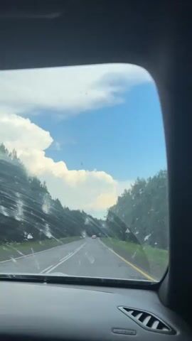 The video, captured from the interior of a vehicle, shows a view through the windshield. The scene is an asphalt road with white lane markings and a yellow line on the right edge, suggesting it is a rural or suburban highway. Trees line both sides of the road, and the sky is a bright blue with large, fluffy white clouds. The windshield appears to have water streaks and a few small debris marks, possibly from bugs or dirt.

In the distance, there are a couple of cars visible on the road. The lighting suggests it is daytime, likely late morning or afternoon. The presence of cumulus clouds and the clear blue sky indicate fair weather, although the streaks on the windshield could imply recent rain or moisture. The lush green foliage of the trees points to a temperate season, possibly summer or late spring.

There is no visible text in the video. The overall mood is peaceful and conveys the experience of a scenic drive. The dynamic element is the motion of the vehicle, indicated by the slight blur and the passage of scenery.