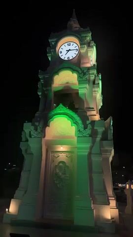 This media file captures a nighttime scene in Surat Thani City Municipality, Thailand. The central subject is a tall, ornate clock tower, illuminated with green and white lights. The clock face displays the time as approximately 9:10. The tower has intricate architectural details, including carvings and a spire. In the foreground, there are decorative white lamp posts, and a bench is visible to the left.

The camera pans to the right, revealing a street scene with buildings on either side. The buildings appear to be shops or businesses, with some showing signs of illumination. Several cars are parked along the street, and a few vehicles are in motion, including what appears to be a motorcycle. The overall setting is urban, with the darkness of night contrasting with the artificial lighting.

The time of day is clearly night, indicated by the dark sky and the prominent artificial lighting. There are no discernible weather conditions. The presence of illuminated buildings and moving vehicles suggests that the area is active at night. The mood is somewhat serene, with the illuminated clock tower being a focal point. No people are clearly visible in detail, though silhouettes might be present within the tower or in the distant buildings. There is no visible text in the media file.