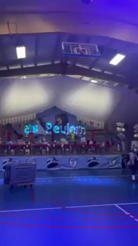The video shows a stage set up in a gymnasium. The stage is decorated for a celebration, possibly a local event. There is a large banner with the words "CS Peulem" in neon lights and an image of a person in historical garb holding a sword or baton. The stage is flanked by colorful decorations and balloons. Tables and chairs are arranged on the stage, and there's a banner along the front with various logos and designs. A person in a jacket and hat is standing to the side of the stage. The gym floor has blue and red lines. A metal cart is on the floor.