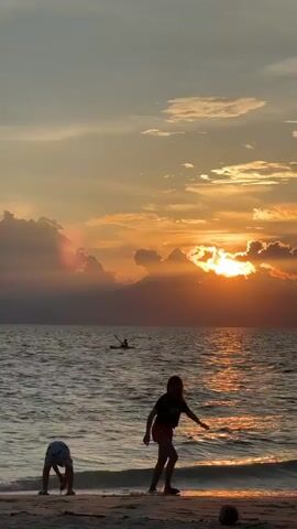 The video shows a serene beach scene at sunset. The sky is ablaze with orange and yellow hues, with clouds casting dramatic shadows and shafts of light. The sun is partially obscured by clouds, creating a beautiful, diffused glow. The ocean is calm, with gentle waves lapping at the shore. In the foreground, two children are playing on the sand. One child, on the left, is bent over, seemingly digging or playing with sand. The other child, on the right, is standing and appears to be interacting with someone off-camera or looking at something in the distance. In the middle ground, a small boat with a person in it is visible on the water, adding a sense of peaceful human presence. The overall atmosphere is tranquil and picturesque, capturing a moment of natural beauty and simple childhood joy.