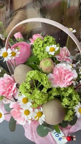 The video showcases a flower shop's display of Easter baskets. There are multiple baskets, each filled with different arrangements of flowers and decorated eggs. The flowers include carnations, daisies, and daffodils, in colors such as pink, white, green, and yellow. The decorated eggs have speckled designs and come in shades of brown, yellow, and green. Price tags are attached to the baskets, displaying the prices. The baskets are arranged on a gray surface, likely a display table in front of a shop window.
