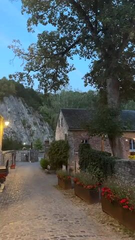 This is a vertical, handheld video of a cobblestone path leading through a village or town. The path is lined with stone walls and planters filled with red flowers. On the right side of the path, there is a stone building with an outdoor seating area illuminated by string lights. Further down the path, there is a gate with a sign and a freestanding sign in front of it. A stone tower with a pointed roof is visible in the background. The sky is a deep blue, suggesting it is dusk or early evening. The environment is lush with trees and greenery on the surrounding hillsides. There are no people visible in the video.