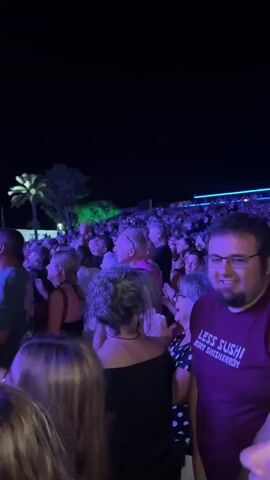 A concert crowd is seen from behind as the audience faces a stage with musicians. The stage is illuminated with bright blue lights. A person in the foreground on the right is wearing a purple t-shirt. In the center of the frame, a person with gray hair is clapping. In the background, a palm tree is visible under green lights. The sky is dark.