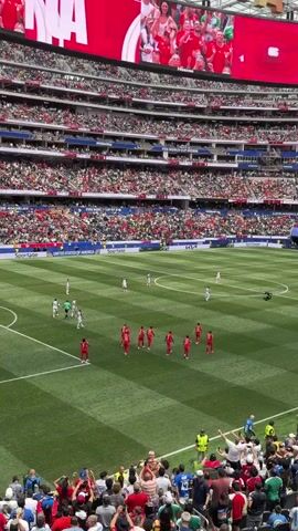 A high-angle, slightly shaky, medium shot captures a wide view of a soccer match in a large stadium. The field is a vibrant green, with white lines clearly marking the boundaries and playing areas. Two teams are present on the field, one in red uniforms and the other in white. The team in red is clustered near the center of the field, facing a group of players in white uniforms who are further down the field. Spectators fill the tiered seating of the stadium, predominantly dressed in red and white, creating a sea of color. A massive screen at the top of the stadium displays a celebratory image of people in red and white, with the word "GOAL" prominently featured. The overall atmosphere appears energetic and enthusiastic, typical of a live sporting event.