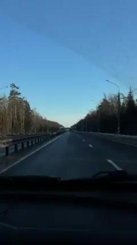 The video shows a driver's point of view on a highway. The sky is clear and blue. On either side of the road, there are trees and patches of snow. Guardrails are visible to the left. Streetlights line the road. A few vehicles can be seen in the distance. The road is wet and black. The car's dashboard is visible at the bottom of the frame.