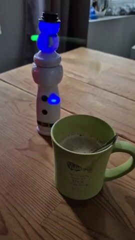 On a wooden table sits a green mug filled with a greenish liquid and a small spoon. Next to the mug is a white snowman-shaped container with three black dots down the front. The snowman is topped with a black hat. All around the scene, and seemingly superimposed onto the image, are a variety of colored lights including red, green, yellow, and blue.
