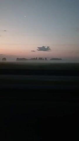 This is a handheld video taken from a vehicle. The scene is a rural landscape at dawn. The sky is a pale pink and blue gradient. A sliver of a crescent moon is visible in the upper left corner. There are a few small clouds. A thin layer of fog or mist hovers over a field of green grass, obscuring the trees and any structures in the distance. The dark foreground suggests the viewer is looking out a window or from a dark interior of the vehicle. A dark strip, possibly a road or the edge of the vehicle's interior, runs horizontally across the bottom of the frame. The video shows a slow, steady movement consistent with a vehicle in motion.