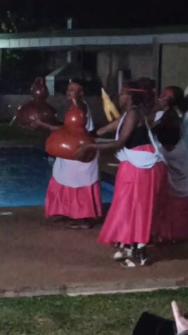 Three women in traditional clothing, including pink skirts, white aprons, and headbands, are dancing outdoors at night near a swimming pool. They are holding large, gourd-shaped objects. The background shows a building and some green space. The dancers are smiling and appear to be enjoying themselves. The scene is lit by artificial lighting.