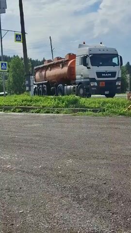 A large white and orange fuel tanker truck is parked on a paved surface. The truck is facing right and is partially obscured by green grass and a yellow barrier. The sky is partly cloudy. There are traffic signs and poles visible in the background. The truck appears to be stationary.