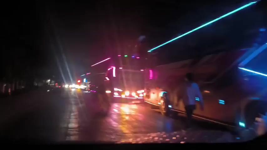 The video shows a nighttime street scene from the perspective of someone in a car, looking out through the windshield.  Several buses are stopped or moving slowly in the same direction as the car. The buses are decorated with bright neon and LED lights, primarily pink and blue. There are other cars visible in the distance, also with headlights on. The road is wet and reflects the light from the buses and cars.