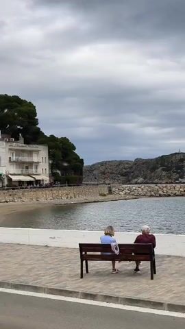 A video shows a coastal scene on a cloudy day. Two people are sitting on a park bench, facing the water. The water is enclosed by a breakwater made of large rocks. Beyond the breakwater, a small, rocky island with a structure on top is visible. The shoreline is lined with a low, white wall. Behind the wall, there's a pedestrian walkway paved with brick. On the walkway, there are lampposts and a wooden structure with a blue and green sign with white writing. The video pans slowly, showing the scene from right to left. The overall impression is of a calm, seaside town.