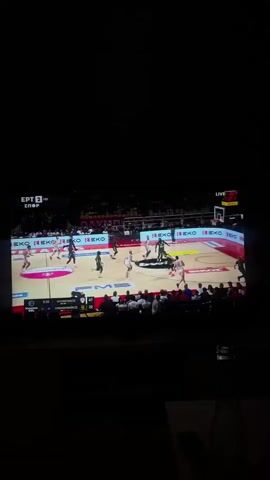 A video captures a live basketball game displayed on a TV screen in a dark room. The television screen shows an indoor basketball court illuminated by arena lights, with players in black and white/light grey uniforms actively engaged in the game. Spectators are visible in the stands around the court.

The scoreboard on the TV screen indicates "8:23" remaining in the game and displays the scores as "24" for "ΟΛΥΜΠΙΑΚΟΣ" (Olympiacos) and "47" for "ΠΑΝΑΘΗΝΑΪΚΟΣ" (Panathinaikos) in the "Q3" (Quarter 3). Various corporate logos are visible on the court and advertising boards, including "bwin," "EKO," "Piraeus," and "NOVA." In the top left corner of the screen, the channel logo "ΕΡΤ 2 HD" and the word "Σπορ" (Sports) are displayed, along with "LIVE" in the top right.

The audio features a male commentator speaking in Greek, describing the ongoing play. The low light in the room suggests the video was recorded during the evening or night. The overall impression is of a sports fan watching a professional basketball match on television.