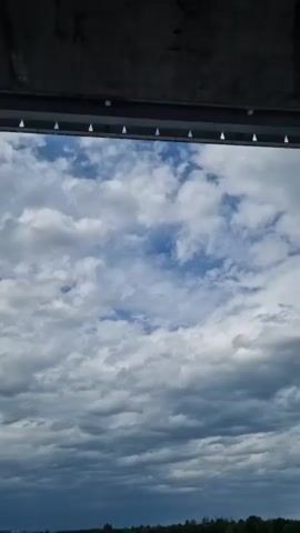 The video captures a low-angle view from underneath a bridge, with the camera moving horizontally from right to left. The underside of the bridge features a grey concrete structure above a long, reddish-brown beam. A yellow diamond-shaped sign is affixed to the reddish-brown beam. Above the bridge structure, a partly cloudy sky with white clouds against a blue background is visible. A railing is briefly seen along the top edge of the bridge. No people are present.