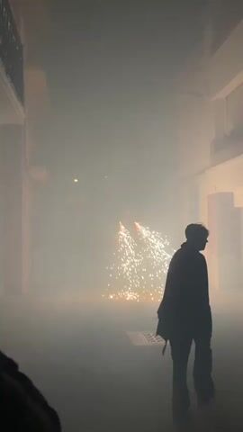 A person holding a phone films a street performance at night. In the foreground, the back of a person wearing a hooded jacket is visible. In the middle ground, another person, silhouetted and wearing a jacket, walks away from the camera. Further in the background, a costumed performer, possibly a dragon, is seen with sparks flying from its mouth. The street is filled with smoke and illuminated by the sparks. The environment appears to be an urban street with buildings on either side.